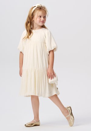 Cremfarbendes Kleid mit Puffärmeln, strukturiertem Stoff und gestuftem Rock. Kombiniert mit goldenen Ballettschuhen und einer kleinen weißen Handtasche.