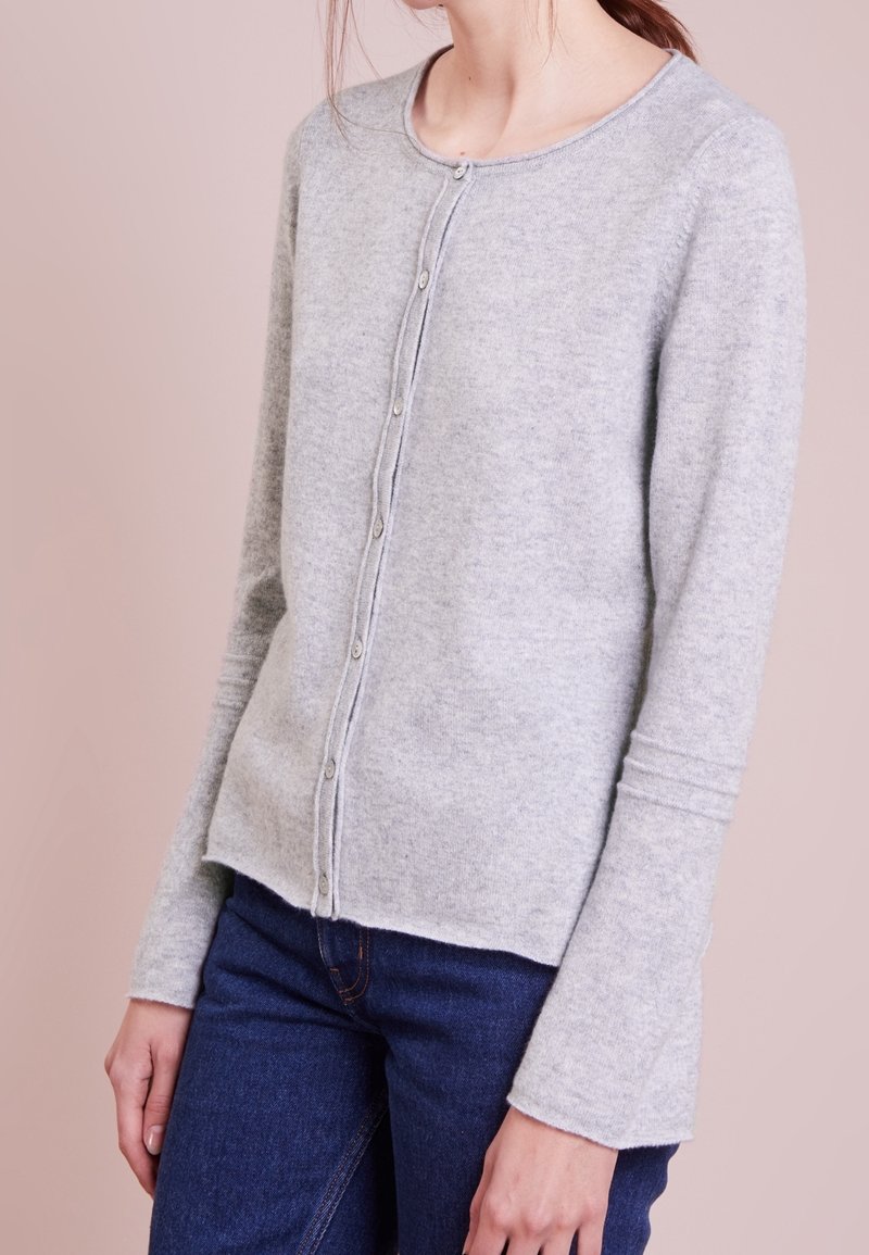 Person, der einen hellgrauen, knöpfbaren Cardigan mit langen Ärmeln und dunkelblauen Jeans trägt, steht vor einem blassen rosa Hintergrund.