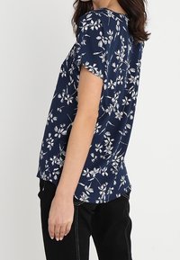 Námořnická modrá blůza s krátkým rukávem a bílým květinovým vzorem, volný střih, měkká tkanina, zaoblený výstřih, viditelné švy, nošena s černými kalhotami.