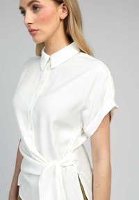 Femme blonde portant une blouse blanche à manches courtes avec un col et une taille nouée, sur un fond gris uni.