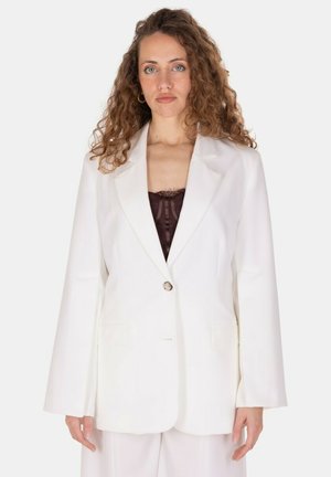 Donna con capelli ricci che indossa un blazer bianco monopetto sopra una maglietta scura, in piedi davanti a uno sfondo bianco uniforme.