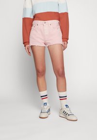 Mujer vistiendo unos pantalones cortos de mezclilla deshilachados en rosa claro, un sudadera de color bloque en blanco, azul y óxido, calcetines blancos con rayas rojas y azules, y zapatillas Adidas blancas.