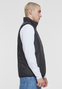 Schwarze gesteppte Weste mit hohem Kragen, Reißverschluss, seitlichen Taschen und glatter Textur, getragen über einem weißen Langarmhemd.