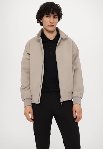 Beige bomberjacka med ståkrage, dragkedja framtill och elastiska ärmslut. Buren över en svart pikétröja, ihop med svarta byxor.