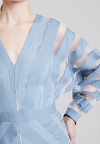 Ljusblå klänning med djup V-ringning, med genomskinliga randiga ärmar och ett bälte i midjan. Den strukturerade tyget kontrasterar med släta detaljer.