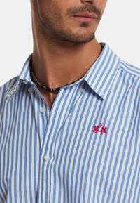 Men's button-up shirt with blue and white stripes, cotton fabric, featuring a small red embroidered logo on the chest and a classic collar.