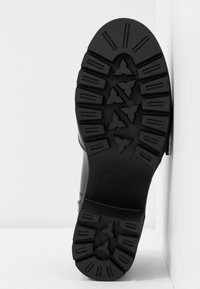 Semelle en caoutchouc noir d'une botte avec des rainures profondes, une finition lisse et un logo en relief, inclinée sur un fond blanc uni.
