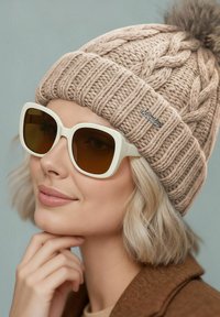 Beige knitted beanie with a pom-pom on top and a silver tag, paired with oversized white sunglasses with brown lenses.