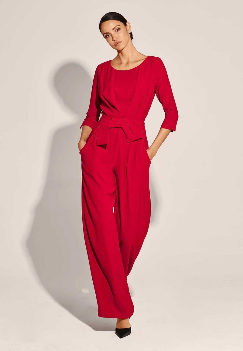 Rode jumpsuit met een riem in de taille, driekwart mouwen, een wijdpijpen ontwerp en zijzakken. Smakkelijk stof, minimalistische snit.