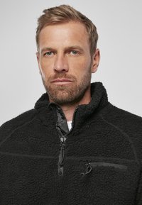Pullover in pile nero con colletto alto, chiusura frontale a mezza zip e tasca sul petto con zip. Presenta un materiale testurizzato e una fodera interna liscia.