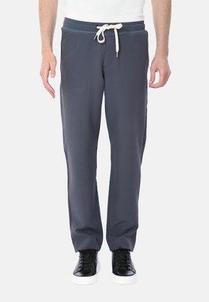Uomo in pantaloni grigi scuri con cordoncino e sneakers nere, in piedi su uno sfondo bianco semplice.