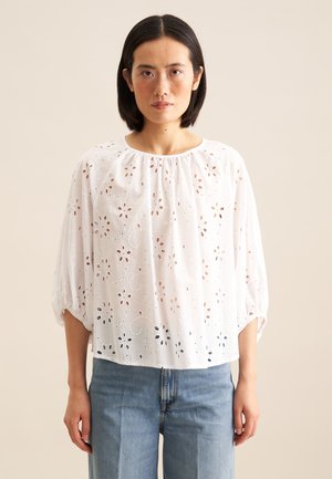 Blusa bianca di cotone con decorazioni floreali a occhiello, scollatura rotonda e maniche a tre quarti, abbinata a jeans di denim blu chiaro.