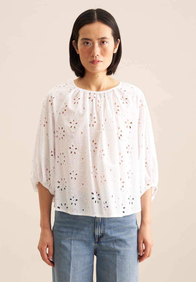 Hvid bomuldsbluse med blomster-øjelet design, rund halsudskæring og trekvartærmer, parret med lyseblå denim jeans.