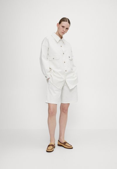 Camicia in denim bianco con due tasche frontali, abbinata a pantaloncini bianchi. Scarpe slip-on marroni e beige con una cintura decorativa sulla punta.