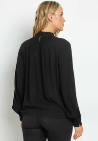 Blusa nera a maniche lunghe con collo alto, chiusura a bottoni sul retro e dettagli arricciati sulle spalle e sui polsini. Tessuto morbido.