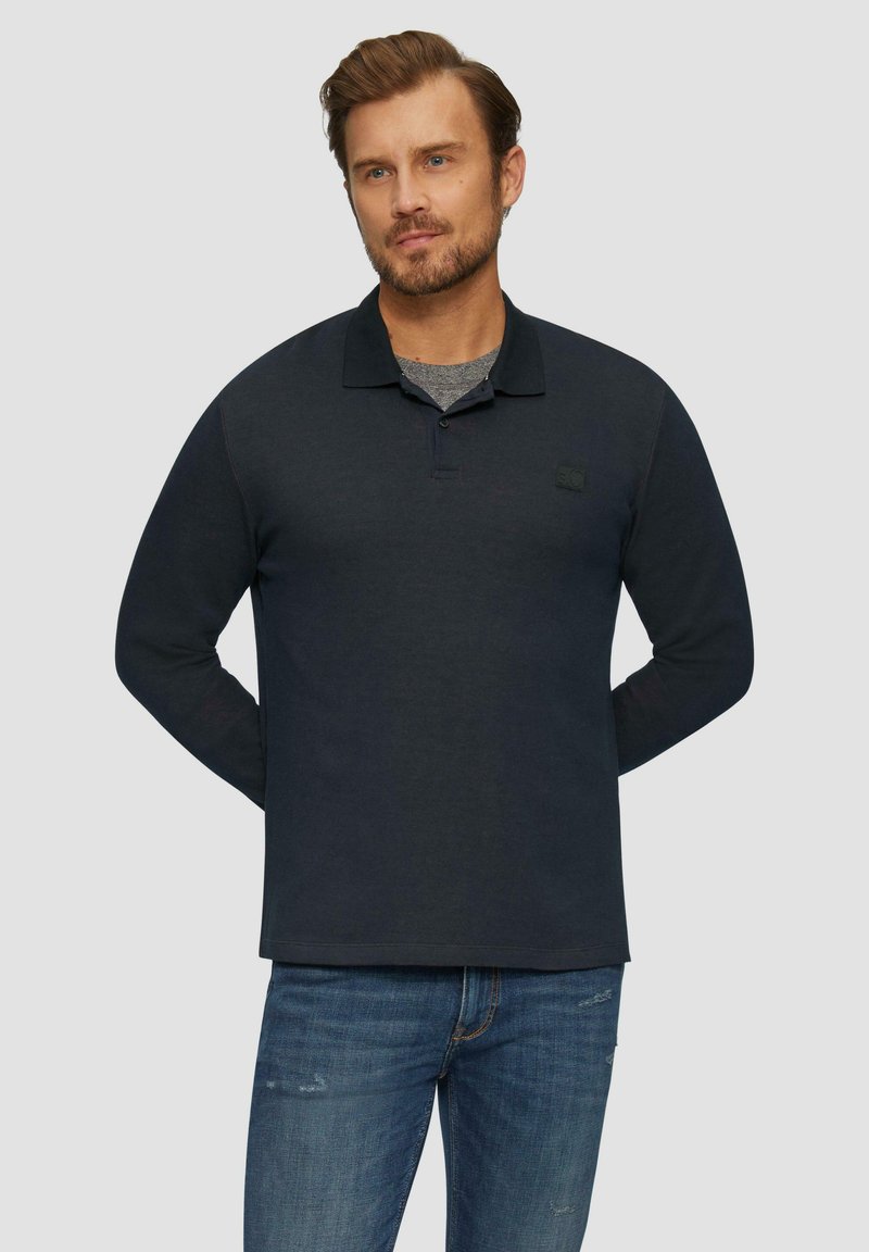 Polo-shirt met lange mouwen in donkerblauwe stof, met een kraag, knoopsluiting en een klein logo op de borst. Gecombineerd met lichtblauwe spijkerbroek.