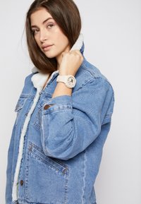 Denim jacket with a fleece-lined collar, blue fabric, button closures, and side pockets. A light-colored wristwatch with a patterned face is worn.