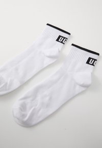 Calze bianche in cotone con un bordo a coste nere che presenta il logo "UG" in bianco. Punta arrotondata e texture morbida, adatte per un uso casual.