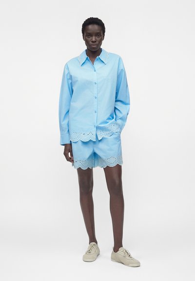 Γυναίκα στέκεται φορώντας ένα ανοιχτό μπλε πουκάμισο με κουμπιά και ταιριαστό σορτς με κυματιστό δαντελένιο τελείωμα, σε συνδυασμό με μπεζ αθλητικά παπούτσια σε λευκό φόντο.