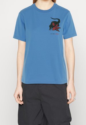 Niebieski t-shirt z krótkim rękawem z czarno-czerwonym nadrukiem stworzenia z twarzą w kształcie kwiatu, noszony z czarnymi szortami.