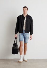 Casaco bomber preto, camisa polo branca, calções de denim claros e sapatilhas brancas. Modelo segura uma bolsa preta com um design texturizado.