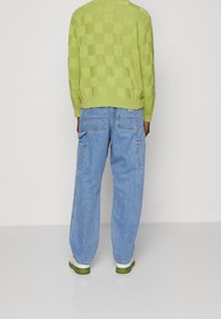 Groen geruite gebreide trui, blauwe losse jeans en witte schoenen met groene zolen. Beide kledingstukken hebben een relaxte pasvorm en een casual ontwerp.
