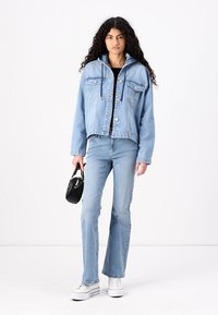 Svetlo modra jeans jakna s kapuco, sprednjimi žepi in gumbi. V kombinaciji s svetlo modrimi širokimi hlačami in belimi športnimi čevlji. Vključena črna torbica.
