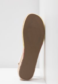Semelle d'une sandale taille 37 avec une semelle plate en caoutchouc marron texturé et un bord tressé, inclinée contre un mur blanc.