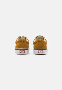 Brune semska sneakers med hvit såle, med polstret krage og et karakteristisk rødt og hvitt Vans-logo på hælen.