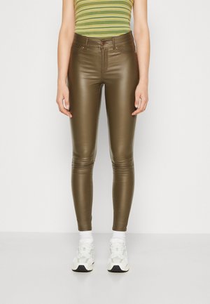 Personne portant un pantalon en cuir vert olive, des baskets blanches, des chaussettes blanches et un top rayé vert sans manches, debout devant un fond uni.