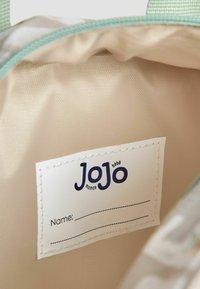 Indersiden af en taske i lys beige med en struktureret overflade, der har et hvidt mærke med brandnavnet "JoJo" og et tomt navnefelt.
