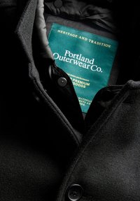 Közelkép egy fekete kabátról, amelyen látható a zöld Portland Outerwear Co. címke arany felirattal a gallér belsejében, valamint az elején látható gomb.