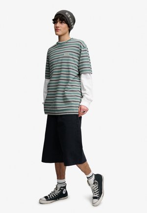Ung person iført en ternet Bench t-shirt over en hvid langærmet skjorte, sorte knælange shorts, sorte sneakers og en grå hue.