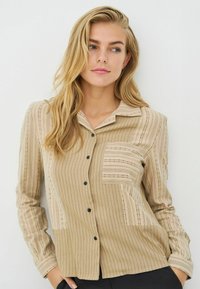 Camicia beige a righe con bottoni e colletto, dotata di taschino applicato e bottoni neri. Tessuto strutturato con linee verticali.