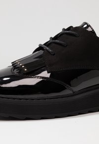 Chaussure en cuir verni noir avec un dessus en daim noir mat, présentant un frange décorative et des clous en métal à l'avant, ainsi qu'une épaisse semelle en caoutchouc.