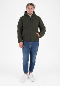Olijfgroene gewatteerde jas met een capuchon, een vooraanlopende rits en zijzakken, gecombineerd met denim jeans en witte sneakers.