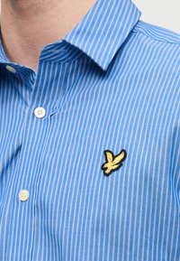 Ανοιχτό μπλε ριγέ πουκάμισο από βαμβάκι με γιακά, διακοσμημένο με μαύρο και κίτρινο κεντητό λογότυπο στο στήθος.