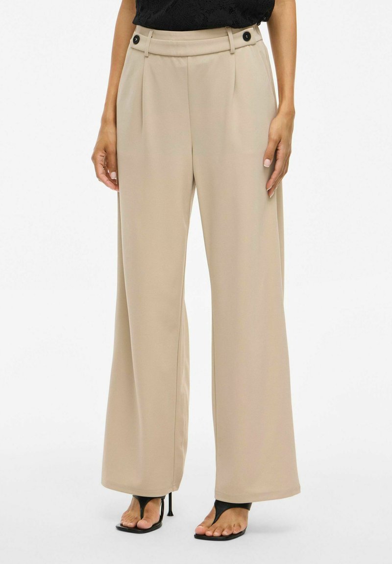 Donna che indossa pantaloni a vita alta beige a gamba larga con dettagli in bottone neri, abbinati a sandali neri con tacco alto e punta aperta.