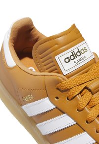 Κοντινό πλάνο ενός ταν Adidas Samba αθλητικού παπουτσιού με λευκές ρίγες, πορτοκαλί κορδόνια και ετικέτα "Samba" στο πλάι και στη γλώσσα.