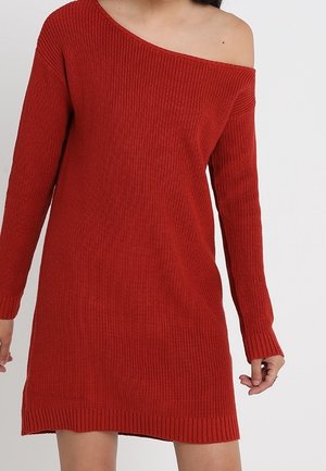 Kvinde iført en langærmet, ribstrikket, rød sweaterkjole med bare skuldre, stående mod en ensfarvet baggrund.