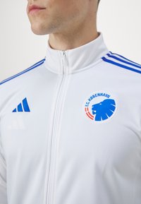 Bijela sportska jakna s patentnim zatvaračem, s plavim prugama i logom koji prikazuje lava. Izrađena od glatke, lagane tkanine, idealna za aktivne aktivnosti.
