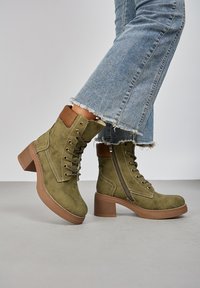 Bottines en daim vert olive avec un talon épais marron, devant à lacets, fermeture éclair sur le côté et détail de col en daim beige, associées à un jean effiloché.