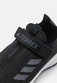 Schwarzer Sportschuh mit einem oberen Teil aus Mesh, verstellbarem Riemen und weiße Sohle. Auffällige graue Akzente und Perforationen für Atmungsaktivität.