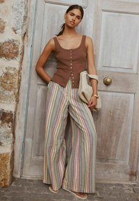 Pruun soonikkoes topp, millel on nööpidega detailid, koos laia värvikirevate triibuliste pükstega. Käes kootud kott, seistes puust ukse ees.