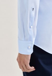 Μανσέτα γαλάζιου πουκαμίσου με λευκά κουμπιά και μικρή μαύρη κεντητή ανθοδέσμη κοντά στη λωρίδα των κουμπιών.