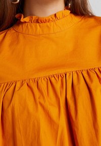 Oranssi puuvillapusero, jossa on korkea röyhelökaulus ja kerätty vartalo. Sujuva pinta, jossa on hillitty kiilto. Ei näkyviä kuvioita tai koristeita.