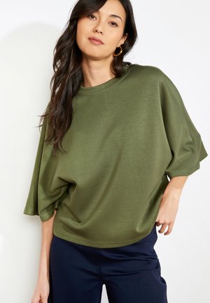 Žena s dlhými tmavými vlasmi, ktorá nosí zlaté kruhové náušnice, olivový nadrozmerný krátky top a tmavomodré nohavice, pózujúca na bielom hladkom pozadí.