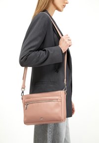 Borsa a tracolla in pelle rosa con tasca anteriore con zip, design slim, tracolla regolabile e dettaglio del logo discreto. Indossata con un blazer grigio.