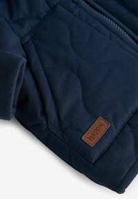 Manche de veste matelassée bleu marine avec poignet côtelé, fermeture éclair et écusson en cuir marron marqué "happy" près du bord de la poche.