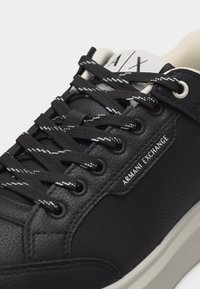 Baskets en cuir noir avec lacets blancs soulignés, "Armani Exchange" imprimé sur le côté, et languette rembourrée blanche avec logo "A|X".